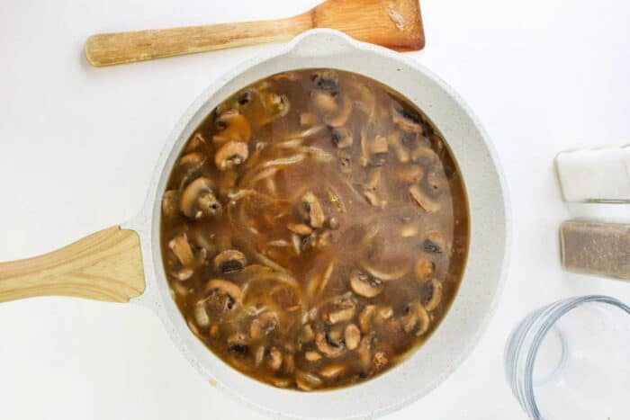 A white skillet filled with savory mushroom and onion gravy for Hamburger Steak, next to a wooden spatula, salt, and pepper shakers.