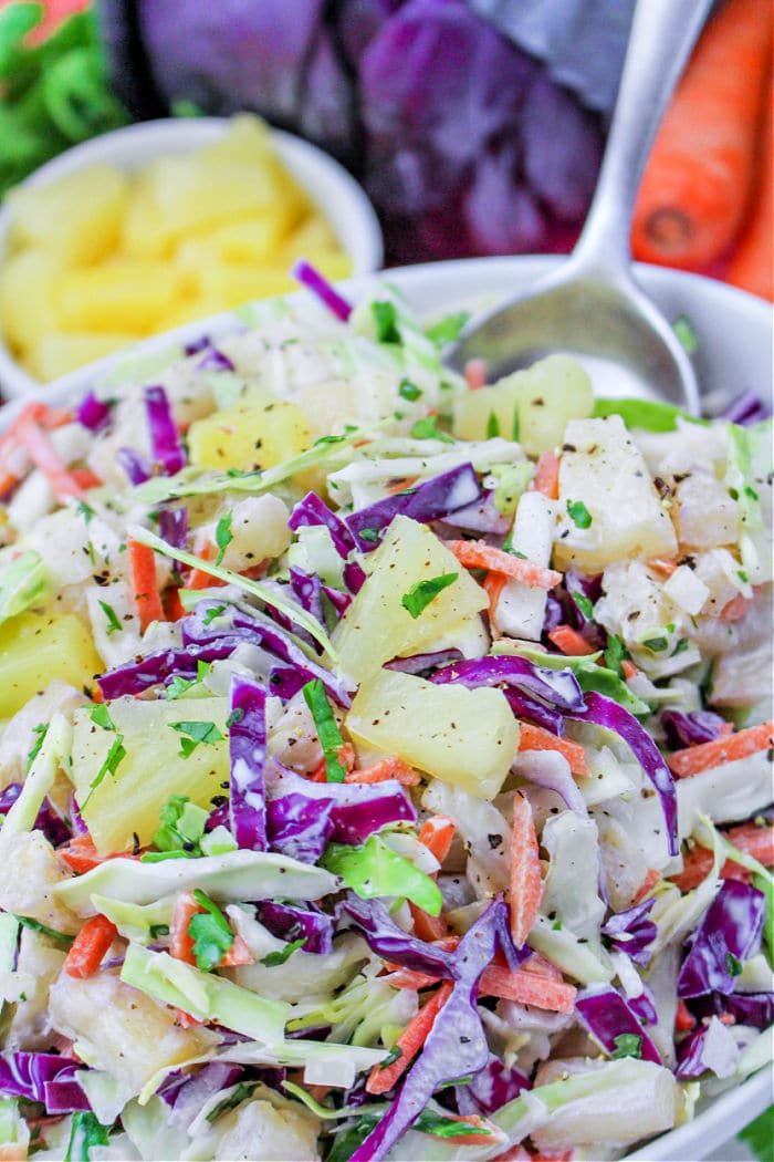 Hawaii coleslaw features colorful cabbage, carrots, and juicy pineapple chunks in a white bowl, sitting beside a spoon and fresh ingredients.