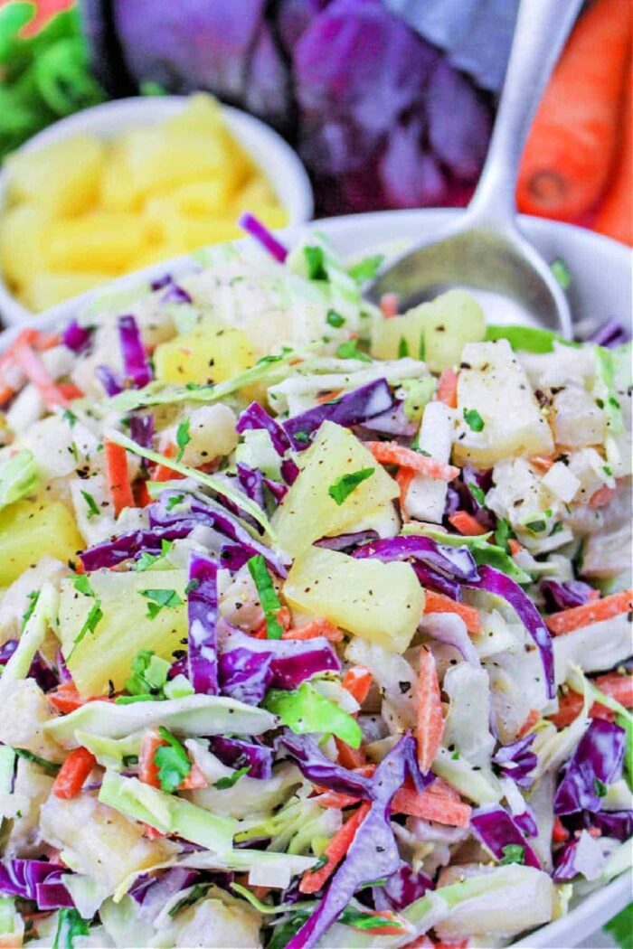 A bowl of Hawaii Coleslaw featuring vibrant purple cabbage, carrots, pineapple chunks, and fresh herbs, with a spoon on the side.