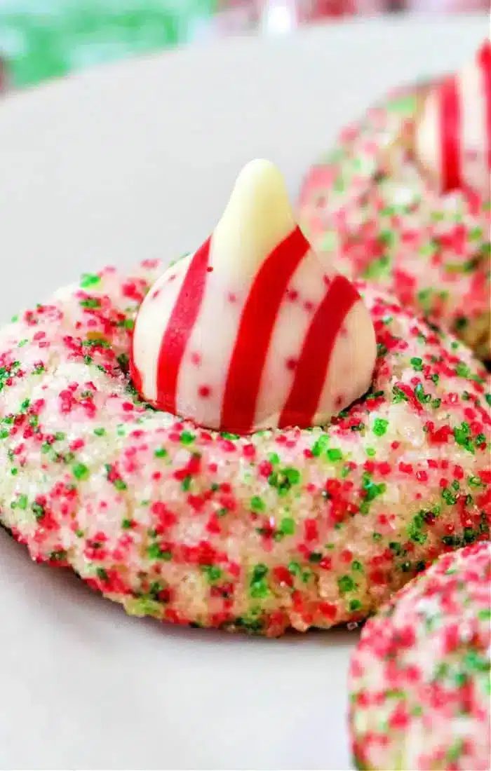 A festive cookie covered in red and green sugar sprinkles features a striped red and white Hershey Kiss shaped like a drop on top. solid for Christmas, this treat sits elegantly on a white plate.