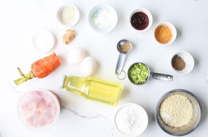 Various ingredients, including eggs, carrot, oil, seasonings, flour, and ground chicken for Homemade Chicken Nuggets with Ground Chicken, arranged on a white countertop.