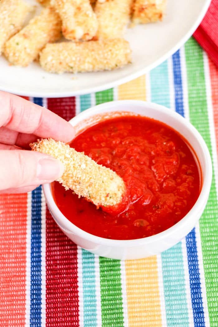 A hand dips a breadcrumb-covered homemade mozzarella stick into a bowl of red marinara sauce. The bowl rests on a colorful striped tablecloth, while more crispy mozzarella sticks, perfected in an air fryer, await on a white plate in the background.