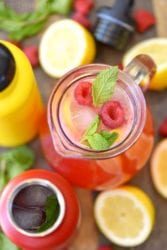 Top view of a pitcher with homemade raspberry lemonade, ice, mint, and lemons surrounded by drink bottles and fresh fruit.