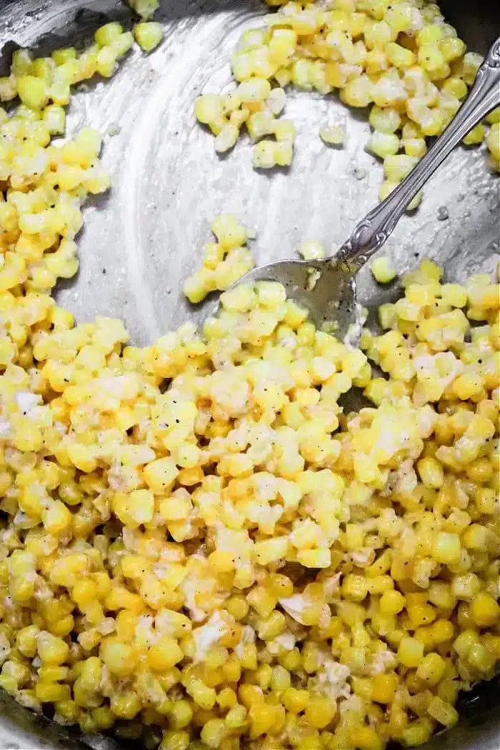 A close-up of a metal dish containing honey butter skillet creamed corn. The yellow kernels are mixed with a creamy sauce, accented by black pepper, while a metal spoon rests in the dish, partially immersed in the corn mixture.