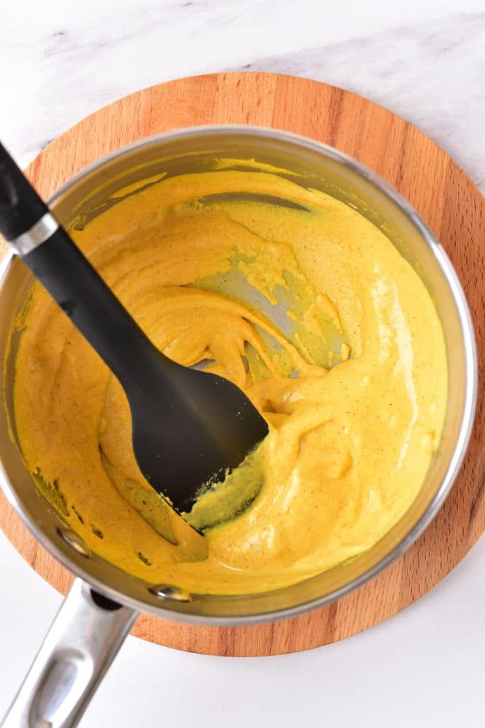 A saucepan filled with creamy hot dog mustard, being stirred by a black spatula on a wooden trivet.