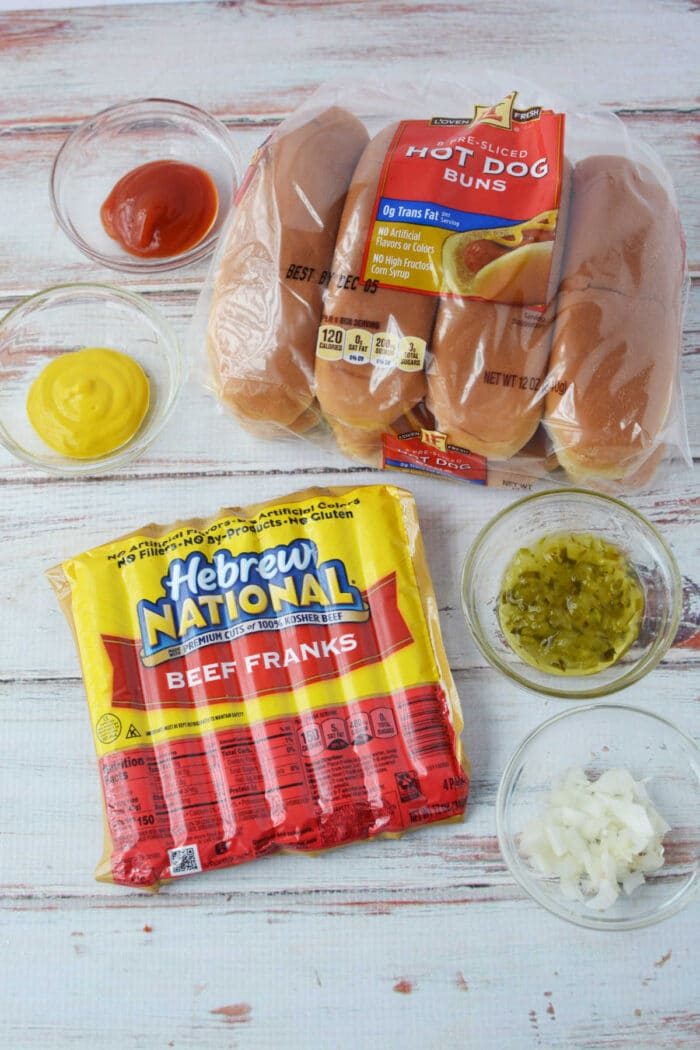 Baked Hot dog ingredients: buns, beef franks, ketchup, mustard, relish, and chopped onions rest on a wooden surface, ready to create solid hot dogs in the oven.