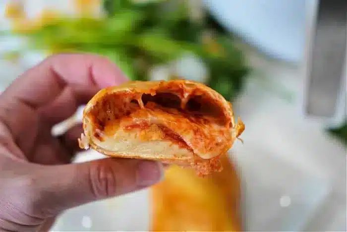 A hand is holding a partially bitten piece of a rolled and baked food item with a tomato sauce filling and melted cheese inside.