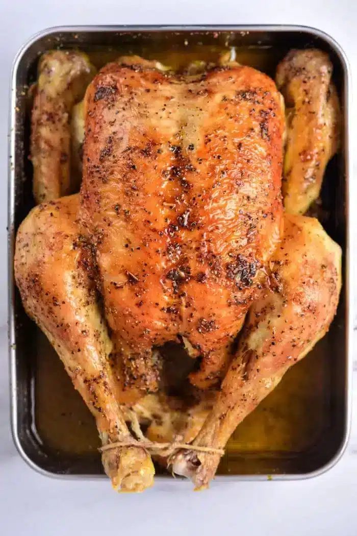 A roasted whole chicken in a baking tray, seasoned with herbs and spices, with tied legs. The skin is golden brown and crispy, resting in a pool of juices. Wondering how long to bake a whole chicken at 350? Ensure solid results by timing your bake for about 1.5 to 2 hours.