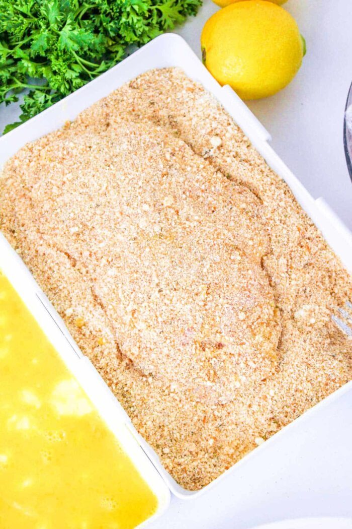 Breaded chicken breast cutlet on a tray, ready to answer the question of how long to bake chicken cutlets, with egg wash and parsley nearby.