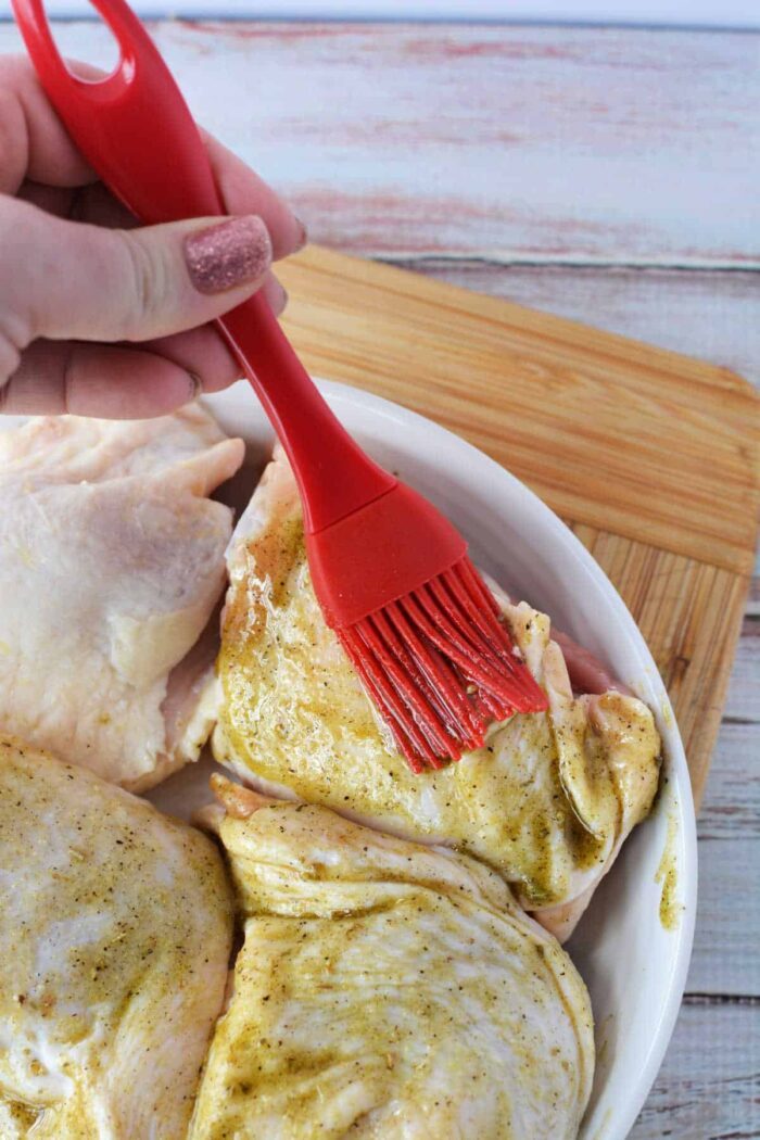 A hand uses a red brush to spread marinade on raw chicken pieces in a white dish, prepping them before discovering how long to bake chicken thighs at 425 for solid results.