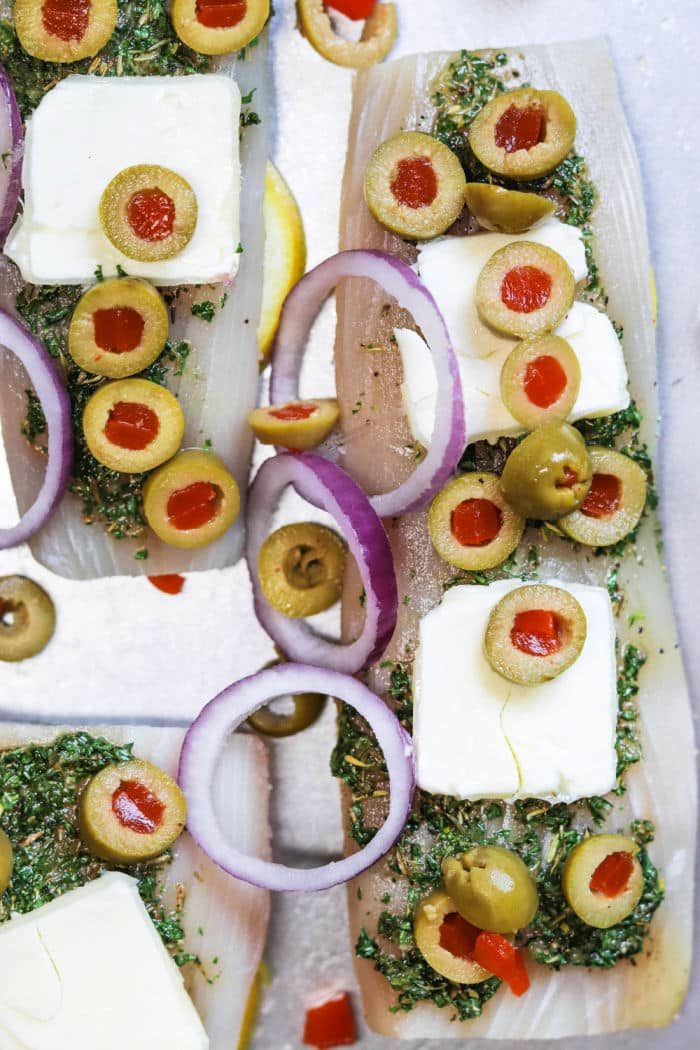 Raw fish fillets topped with herbs, butter, green olives, red onion rings, and pimentos on a tray—solid for trying out how long to bake mahi mahi for tender, good results.