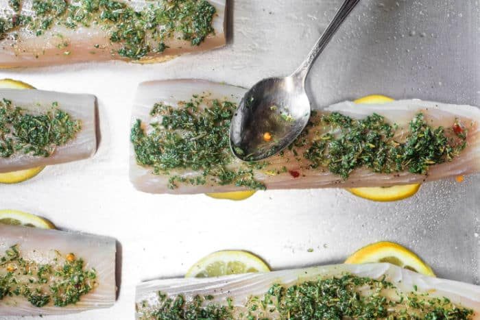 Raw fish fillets on lemon slices with herbs being spread by a spoon, creating a solid pairing while pondering how long to bake mahi mahi for that ideal, flaky texture.