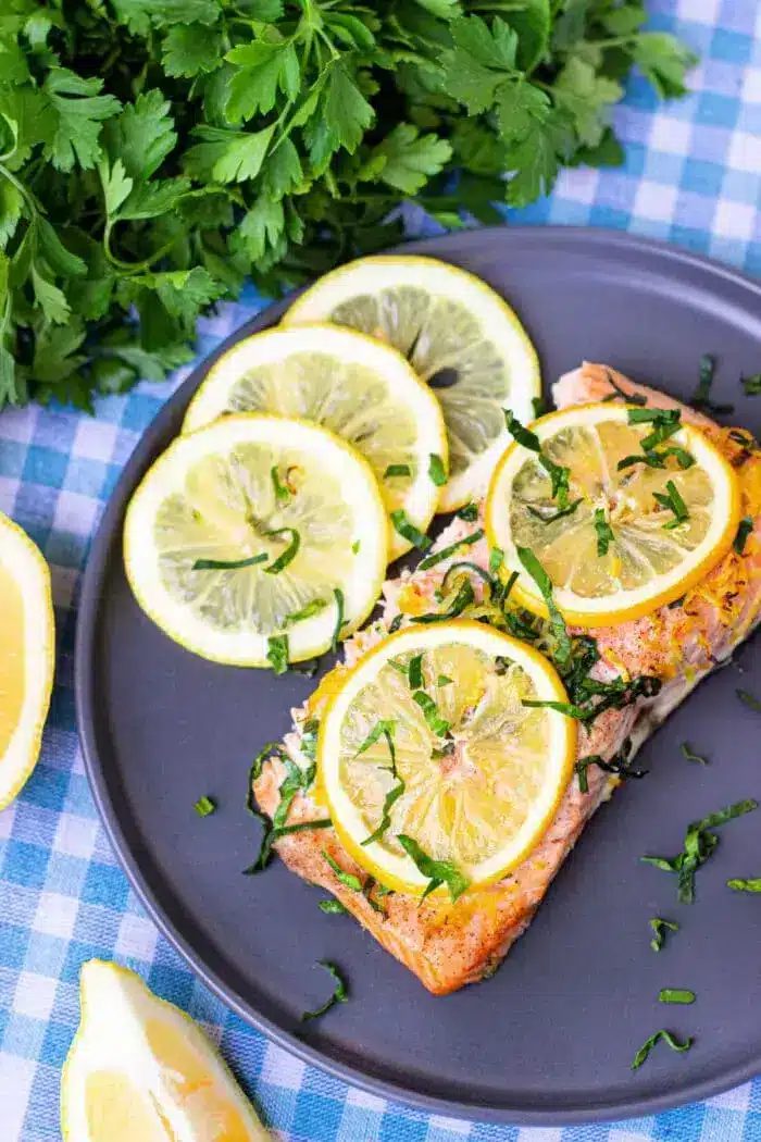 A plate containing a piece of cooked salmon topped with lemon slices and chopped green herbs.