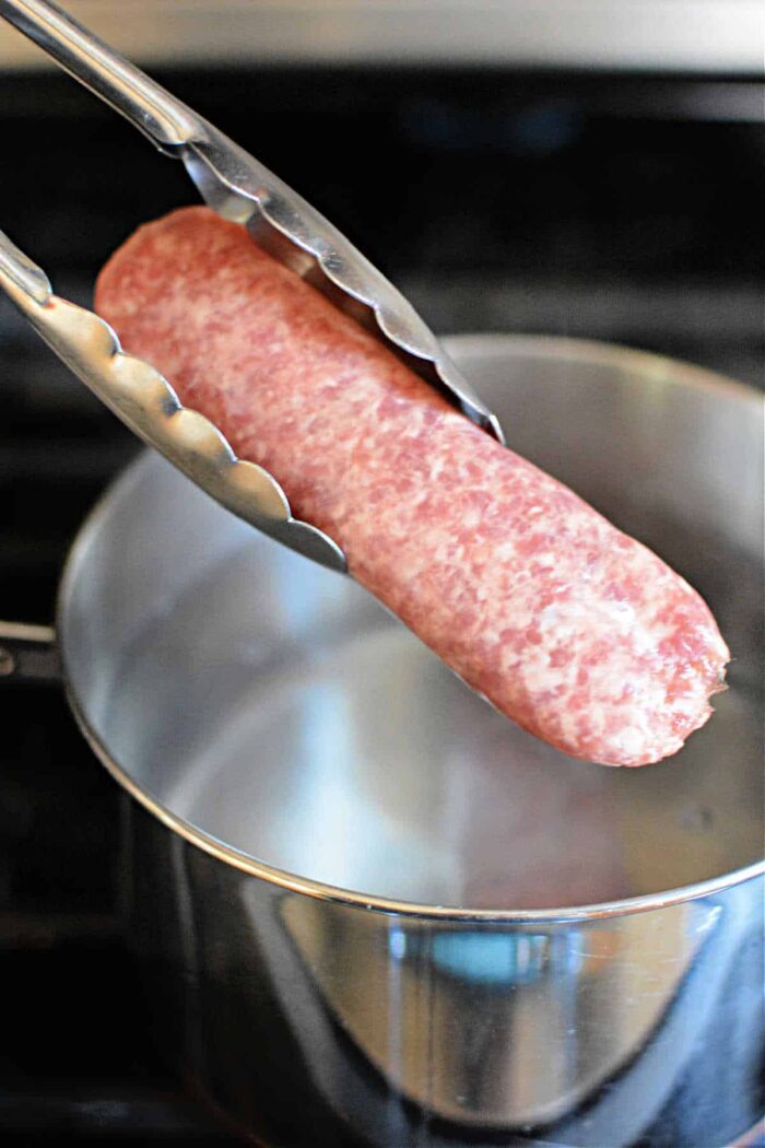 Tongs holding raw brats over a pot of boiling water on the stove, as you ponder how long to boil brats for that solid flavor.