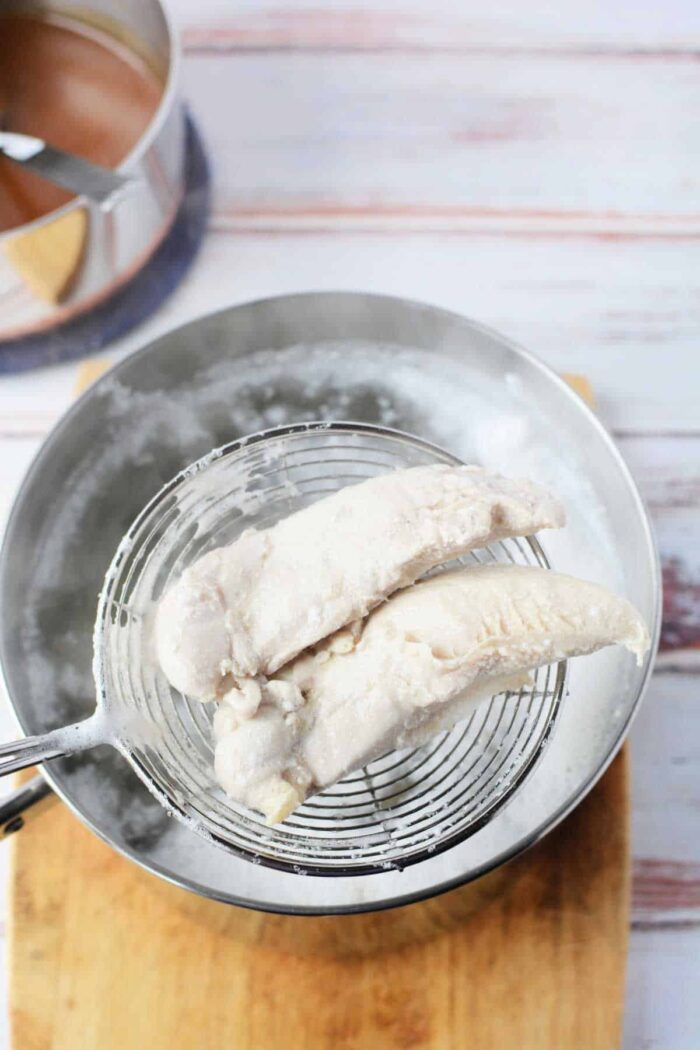 Two cooked chicken tenders, demonstrating how long to boil solid tenders, are being lifted from a pot of boiling water with a wire skimmer and placed on a wooden board.