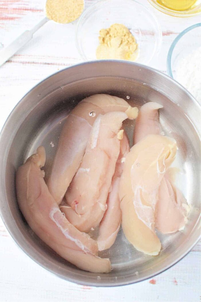 Raw chicken tenders rest in a silver bowl, surrounded by spices and flour on a white surface, evoking the question: how long to boil chicken tenders for tender perfection?.