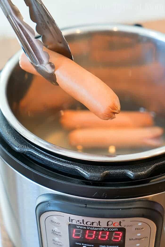 Tongs are holding a hot dog over an open Instant Pot, which is filled with hot dogs. The digital display on the Instant Pot shows a reading of "0003," indicating how long to boil hot dogs for perfectly cooked results every time.