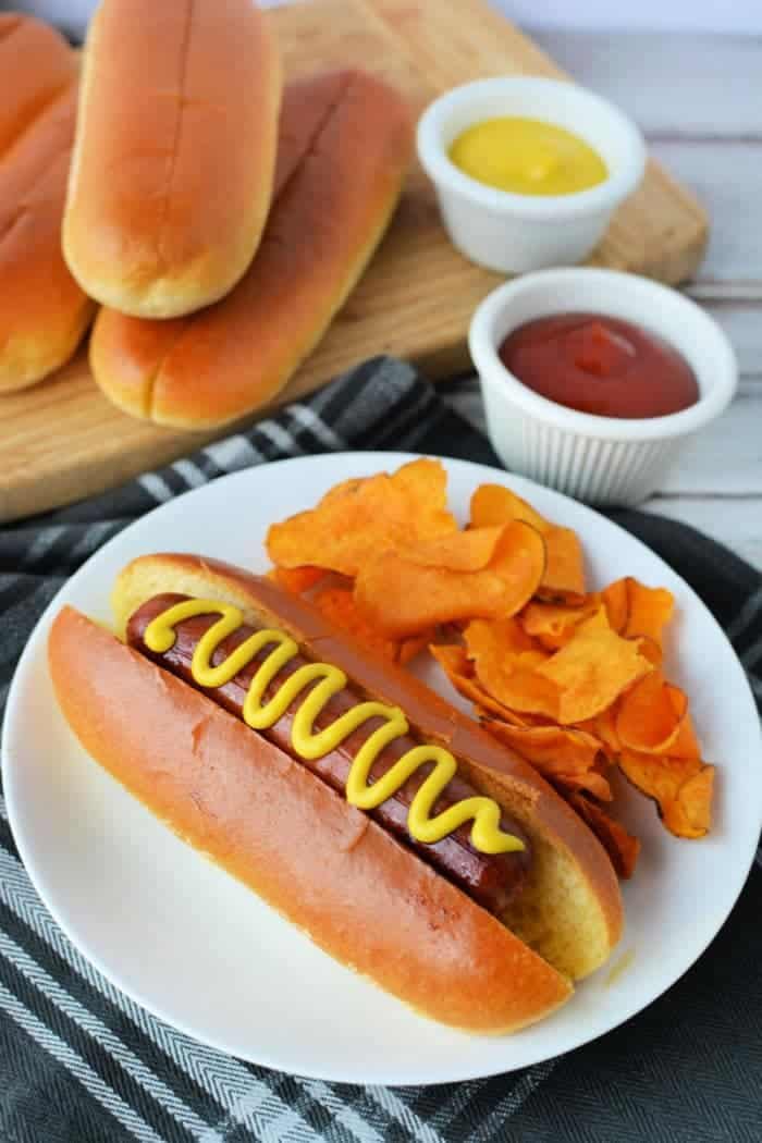 A plate showcases a hot dog in a bun, perfectly boiled to perfection, topped with mustard and accompanied by crispy orange potato chips. In the background, extra buns rest on a wooden board near small bowls of ketchup and mustard—ideal for those wondering how long to boil hot dogs.