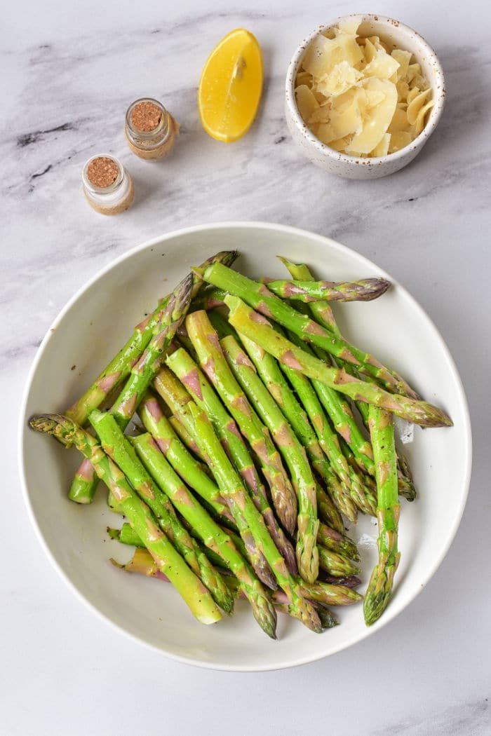 A bowl of asparagus spears with a lemon wedge, shaved cheese, and spices rests on a marble surface, solid for those wondering how long to cook asparagus in the oven for a fragrant delight.