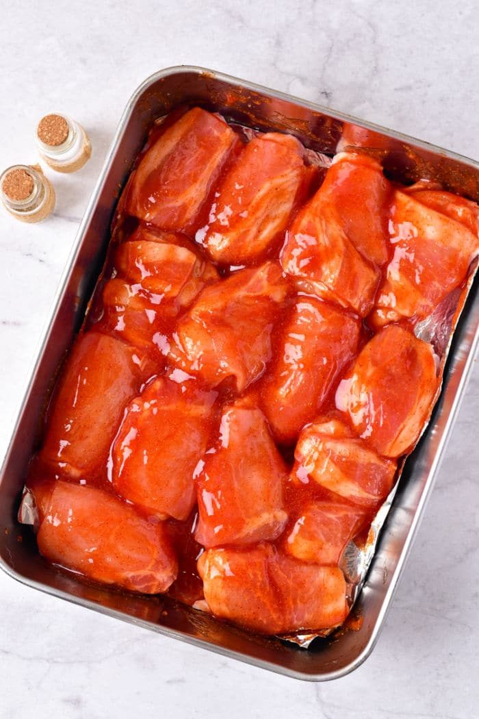 Raw chicken wing pieces coated in red sauce rest invitingly in a baking tray, flanked by two seasoning bottles on the side, ready for transformation. Wondering how long to cook chicken wings in the oven at 375 degrees? This prep sets you up for perfection.