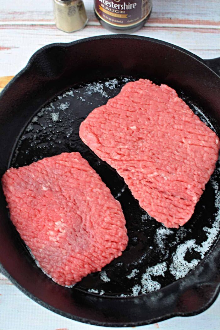 Two raw cube steak patties sizzle in a black cast iron skillet, reminiscent of preparing cube steak; timing is key to achieve solid doneness.