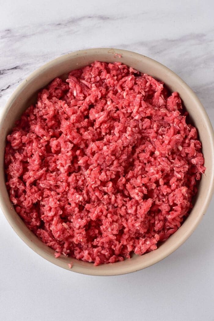 A bowl filled with raw ground meat rests on a marble surface, inviting you to ponder how long to cook ground beef for the solid savory dish.