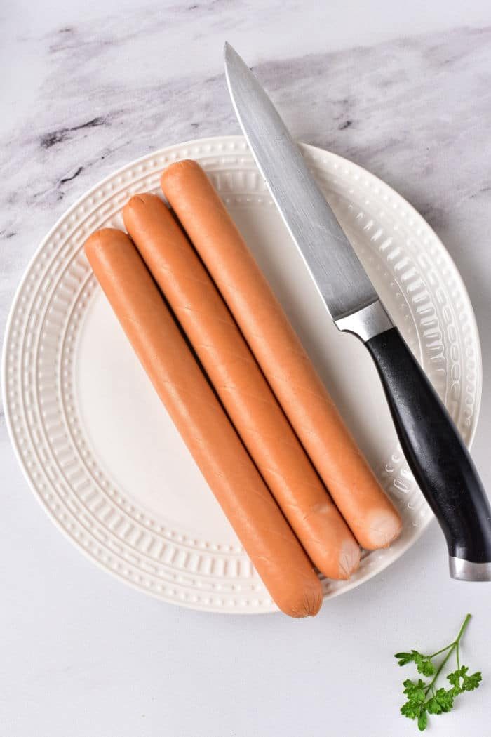 Three hot dogs and a knife rest on a white plate, garnished with parsley. Wondering how long to cook hot dogs in the microwave for that solid texture and taste?