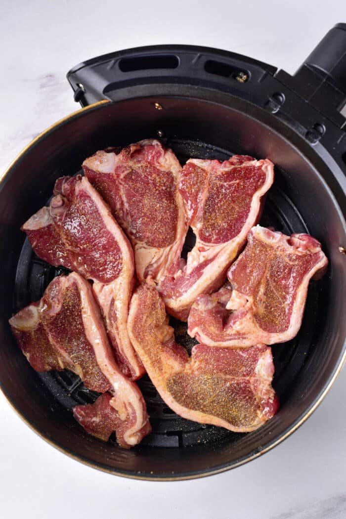 Raw lamb chops seasoned with spices are placed in an air fryer basket, ready for cooking. The basket is open, revealing the marbled meat arranged in a single layer against a light background. Wondering how long to cook lamb chops? Achieve perfection by adjusting the timing according to your desired doneness.