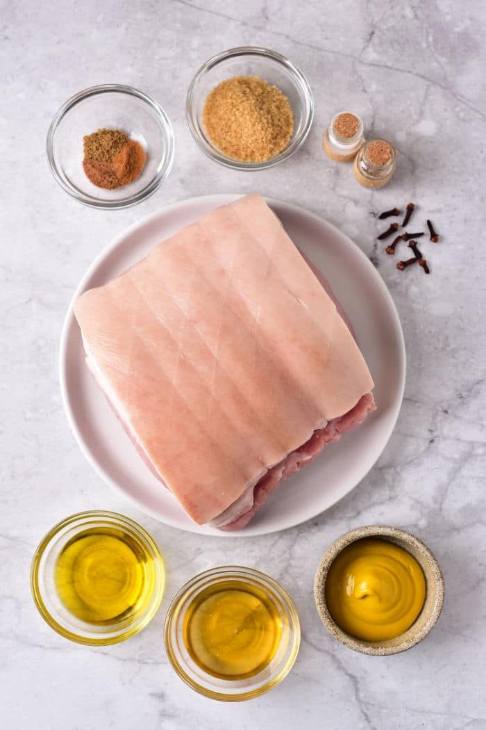 A slab of pork roast on a plate sits elegantly on a marble surface, surrounded by bowls of spices, oil, and mustard, hinting at the solid preparation for those wondering how long to cook a pork roast in the oven at 375.