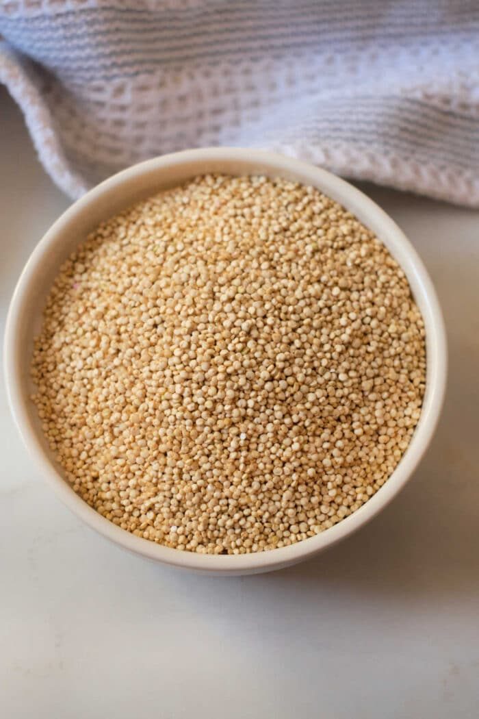 A bowl filled with uncooked quinoa rests on a countertop, accompanied by a textured cloth, all poised to reveal how long to cook quinoa for that solid fluffy texture.