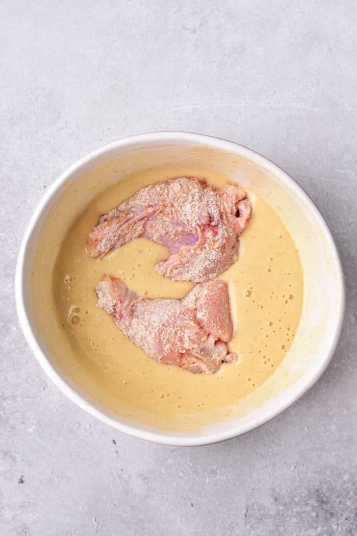 Raw chicken pieces in a creamy tempura batter sit invitingly in a round white bowl on a gray surface, while you ponder how long to deep fry chicken wings for that solid crispiness.