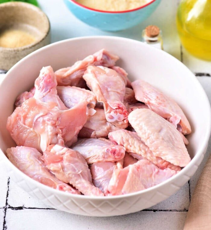 A bowl of raw chicken wings sits on the kitchen counter, surrounded by an array of ingredients, ready to become a flavorful delight. Discover how to bake crispy chicken wings that satisfy your cravings with every crunchy bite.