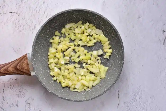 Chopped onions are being sautéed in a non-stick frying pan with a wooden handle on a light textured surface, solid for adding that savory touch to your oven-baked stuffing.