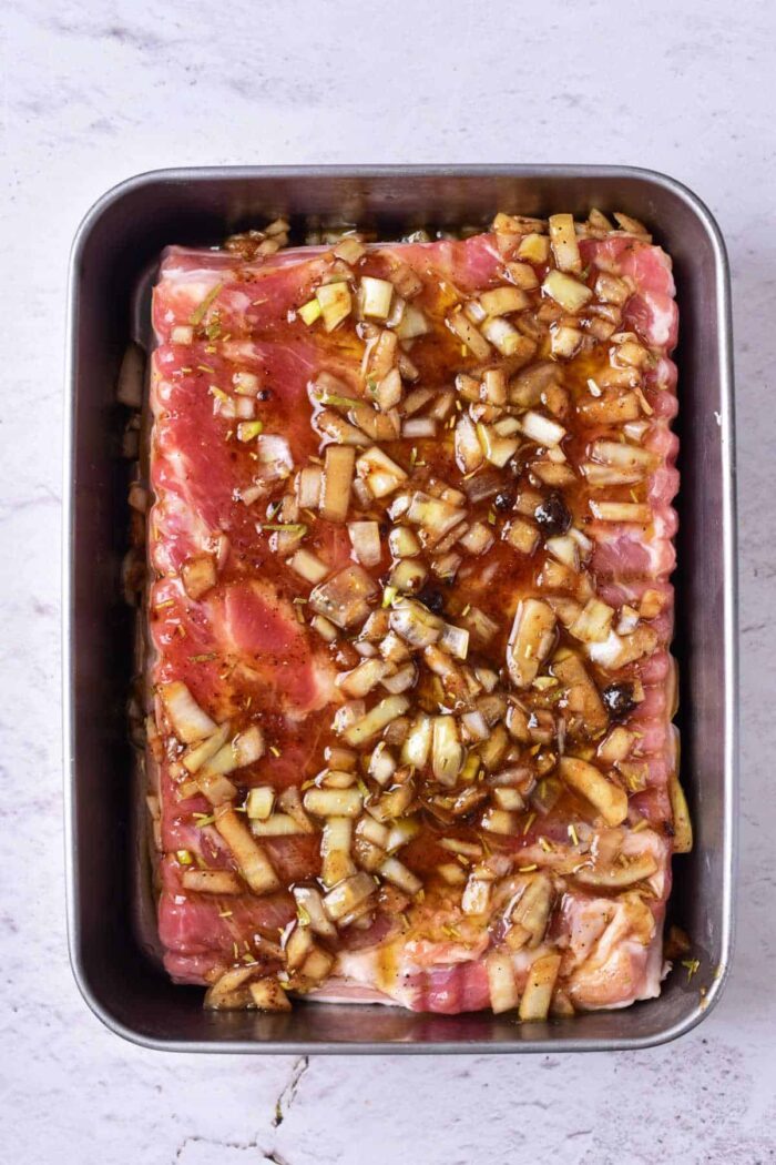 Raw pork belly marinated with chopped onions and sauce rest enticingly in a rectangular metal tray on a light gray surface, evoking the initial steps in learning how to confit pork with good flavor depth.