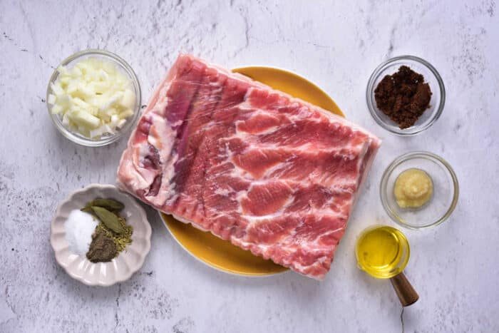Raw pork belly rest on a yellow plate, surrounded by small bowls of spices, chopped onions, and oil on a marble surface—a solid starter kit for those eager to learn how to confit pork.