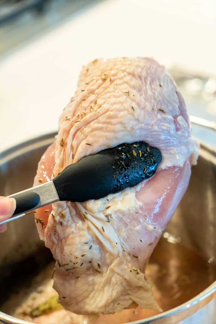 Close-up of a piece of a brined turkey breast, being lifted from a pot with black tongs. The slightly dripping meat hints at marination, ready for cooking. The background features a blurred kitchen counter.