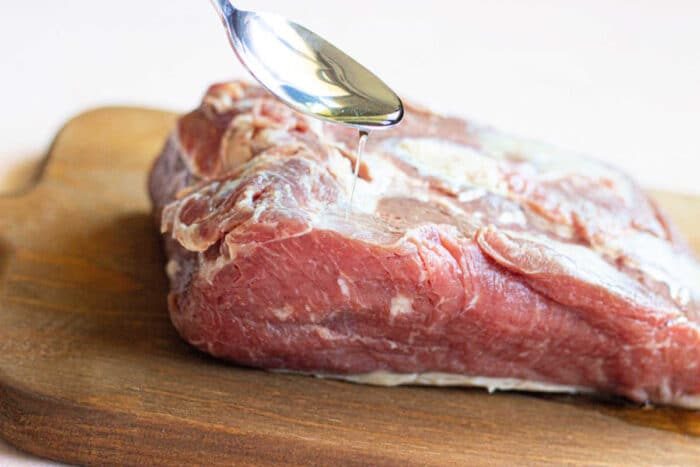 A spoon drizzling oil onto a raw piece of meat on a wooden cutting board sets the stage for mastering how to cook a New York Strip Roast.