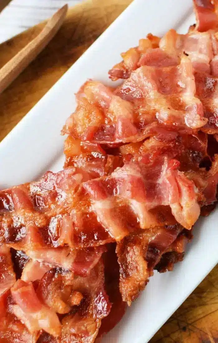 A plate of cooked, crispy bacon strips is arranged on a white rectangular platter. The bacon looks glossy and browned