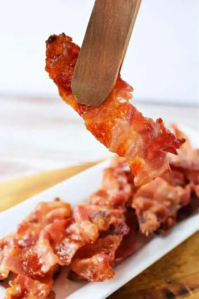 A piece of crispy bacon held by wooden tongs above a white rectangular plate filled with more bacon strips. The background is blurred, and the focus is on the bacon being lifted, showcasing perfectly cooked strips—like what you'd get if you knew how to cook bacon in the oven with aluminum foil