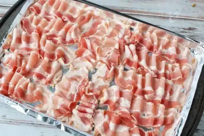 A baking tray lined with aluminum foil is covered with neatly arranged strips of raw bacon, ready to be cooked in the oven.