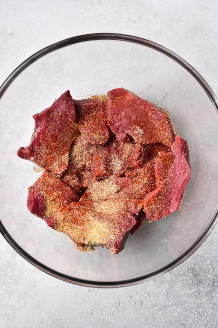 A glass bowl containing raw beef heart pieces all coated with various seasonings, is placed on a light gray surface.
