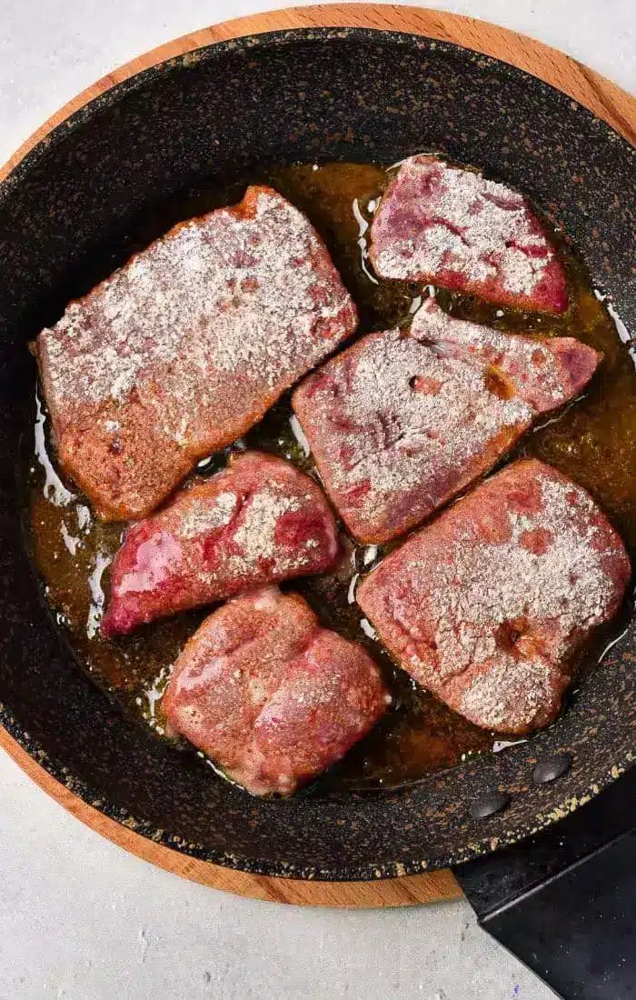 A frying pan with six pieces of liver being cooked in oil. The liver pieces appear to be seasoned with a coating of flour, illustrating how to cook beef liver perfectly.