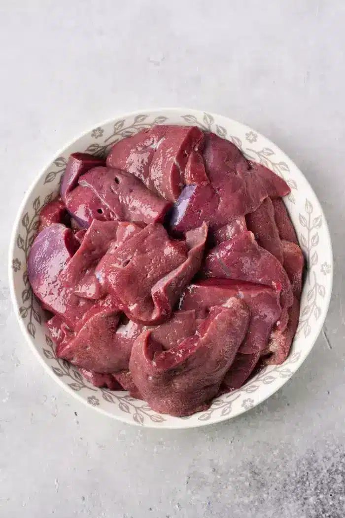 A white bowl filled with raw, sliced chicken liver sits on a light gray countertop.