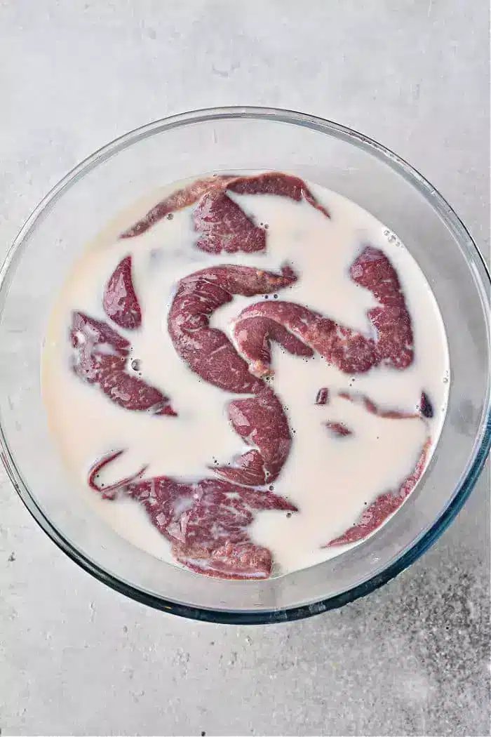 A glass bowl filled with pieces of raw liver soaking in milk rests on a light-colored, textured surface.