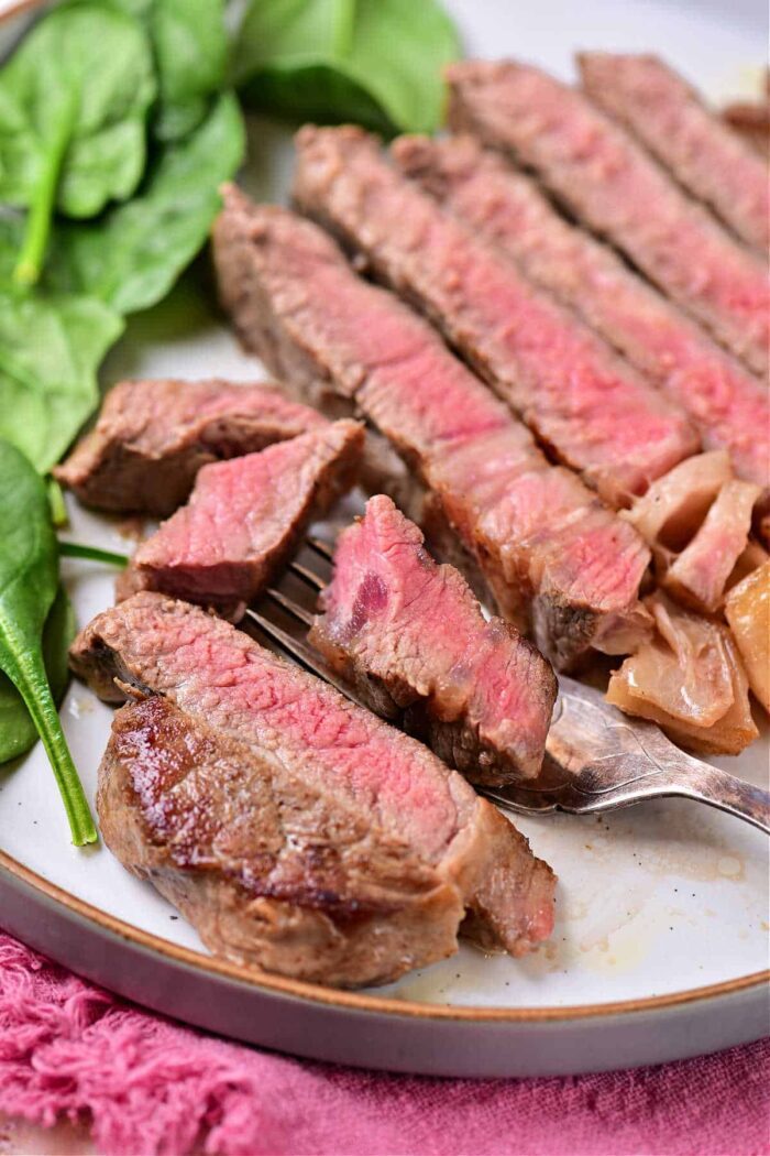 A plate featuring sliced medium-rare steak with visible pink centers is a solid example of how to cook beef strip loin steaks. The steak is placed next to a leafy green, possibly spinach. A fork holds a piece of steak in front of the slices, with a pink cloth in the background adding to the presentation.