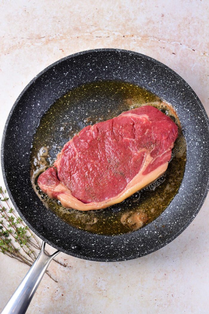 A raw steak, solid for those learning how to cook beef strip loin steaks, is shown lying in a frying pan with some oil. The surrounding surface is a light, textured material, and a small bunch of herbs, possibly thyme, lies next to the pan. The steak appears to be in the early stages of cooking.