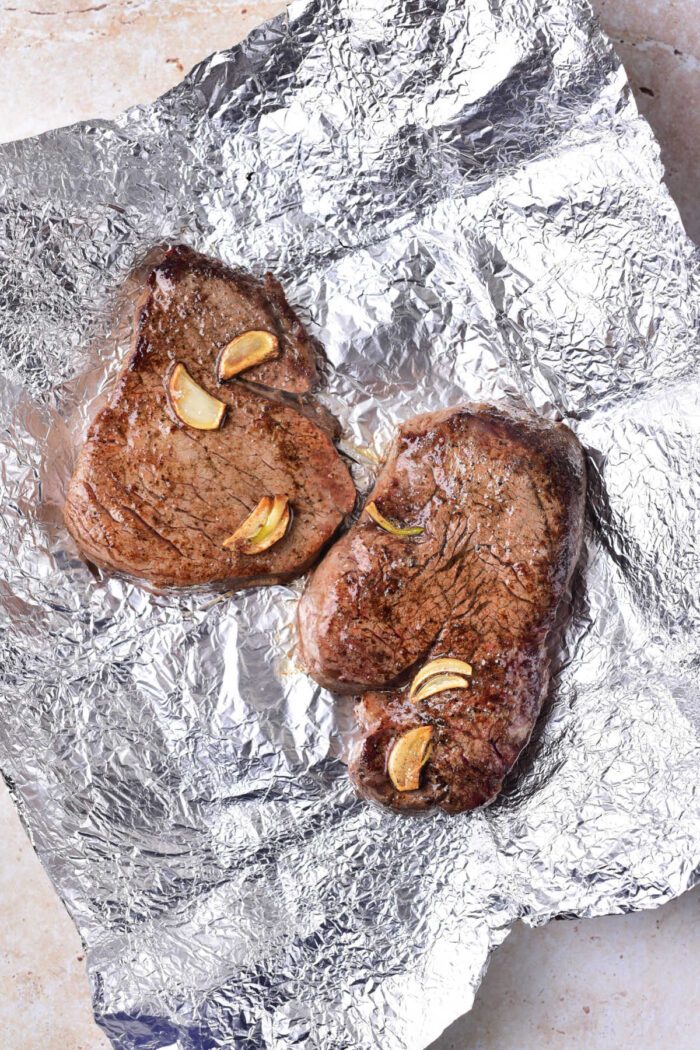 Two succulent cooked beef tenderloin medallions, rest on foil adorned with delicate garlic slices.