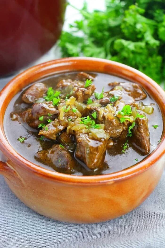 Brown ceramic bowl of beef stew garnished with chopped parsley, placed on a light cloth, with herbs in the background—a good result when you learn how to cook beef tips perfectly.