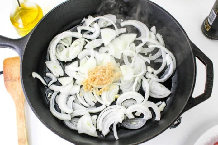 A cast iron skillet on a stovetop containing sliced onions and minced garlic, with steam rising from the pan. A wooden spatula is positioned next to the skillet, and a bottle of olive oil is visible in the background, setting the solid stage for how to cook bratwurst to perfection.