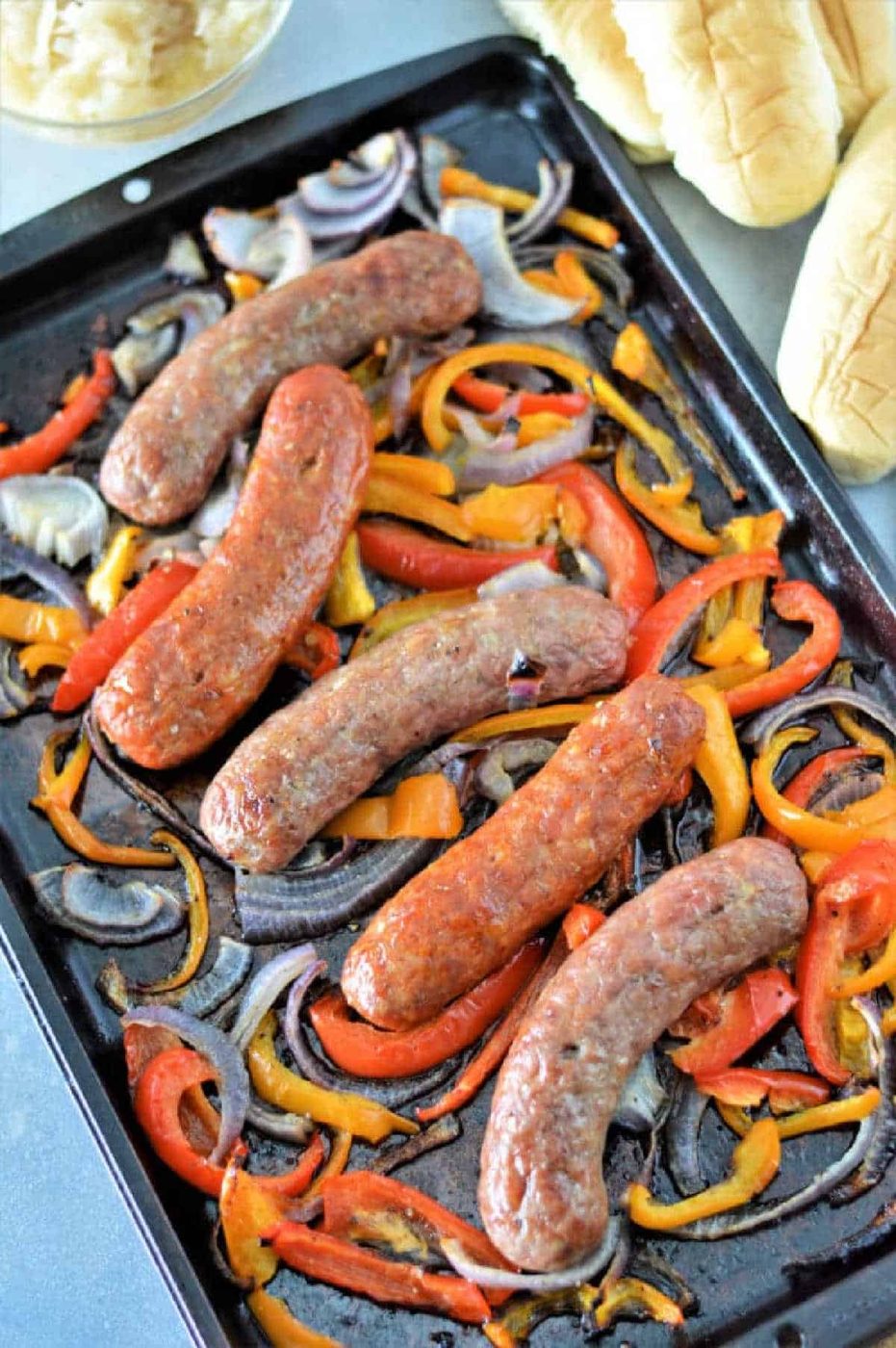 Sausages baked with sliced red onions and bell peppers on a sheet pan—an easy method if you're wondering how to cook bratwurst in the oven, served with hot dog buns nearby.